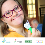 Uśmiechnięta dziewczyna z zespołem Downa trzyma w ręku ozdobioną ręcznie małą latareczkę. Na dole zdjęcia znajdują się logotypy Fundacji Ewangelickie Centrum Diakonii i Edukacji, Państwowego Funduszu Rehabilitacji Osób Niepełnosprawnych, Miejskiego Ośrodka Pomocy Społecznej we Wrocławiu i Biura Bez Barier.