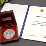 Na stole w otwartym drewnianym pudełku leży medal z wygrawerowanym obrazkiem Wrocławskiego Rynku i napisem Merito de Wratislavia - Zasłużony dla Wrocławia. Medal leży obok dokumentu potwierdzającego nadanie odznaczenia opatrzonego herbem Wrocławia z podpisem Prezydenta Wrocławia.